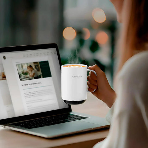 Smart Self Heating Coffee Mug With AI Temp Control And 20 Oz Capacity being held next to a laptop with steam rising from the drink.