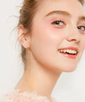 Smiling person with fair skin and light pink makeup, wearing a soft pink top, looking over shoulder against a white background.