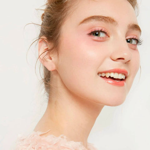 Smiling person with fair skin and light pink makeup, wearing a soft pink top, looking over shoulder against a white background.