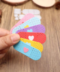 Colorful bandage-shaped card embellishments with heart designs held by a hand, used for crafts and DIY projects.