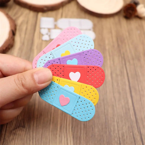 Colorful bandage-shaped card embellishments with heart designs held by a hand, used for crafts and DIY projects.