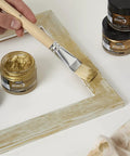 Front view of four Pebeo gilding wax jars (King Gold) with a brush applying gold paste to a wooden frame.