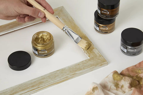 Front view of four Pebeo gilding wax jars (King Gold) with a brush applying gold paste to a wooden frame.