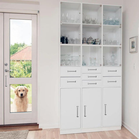 A tall white cabinet with glass doors and drawers in a bright room, featuring metal pull handles, viewed with a dog outside a glass door.