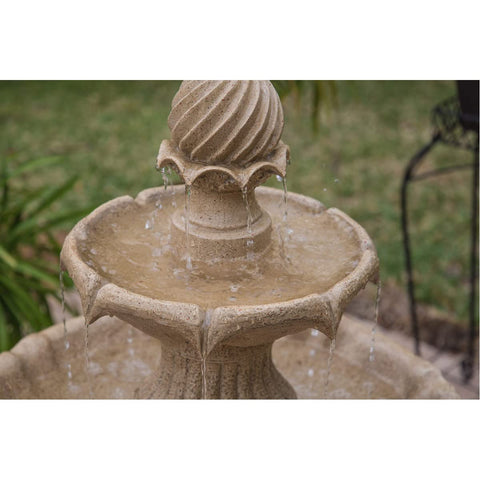 Close-up of a beige stone fountain with water flowing, featuring a spiral pattern top, set in a grassy garden background.