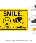 Two Pack Smile Youre On Camera Sign For Outdoor Video Surveillance Signs UV Printed Aluminum 10 By 7 Inches Rust Free For Home And Biz Driveway CCTV Security Signage with smiley face and camera graphics, pre-drilled holes, and UV protection details visible.