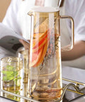 A Game Changer High Borosilicate Glass Pitcher With Secure Tight Lid And Long Mixing Spoon One Point Six Liter Heat Resistant Beverage Carafe pouring iced tea with lemon.