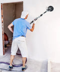 Man using Powerful 710W Drywall Sander With LED Light And Variable Speed 800 To 1800 RPM on a wall. Focus on tool and dust hose.