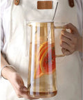 A Game Changer High Borosilicate Glass Pitcher With Secure Tight Lid And Long Mixing Spoon One Point Six Liter heat resistant beverage carafe holding iced tea and fruit slices.