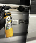 Gloved hand holding a can of T40 Beefed Up Rust Protection And Lubricant For Metal Surfaces near a vehicle hinge.