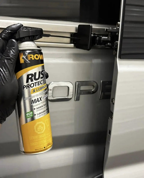 Gloved hand holding a can of T40 Beefed Up Rust Protection And Lubricant For Metal Surfaces near a vehicle hinge.