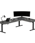 Level Up Your Grind With This Electric Height Adjustable L Shaped Desk Offering Smooth Lift Mechanics And Solid Steel Base, 88 Inch By 55 Inch Corner Workstation, Black Frame Gray Oak Top with laptop and monitor setup.