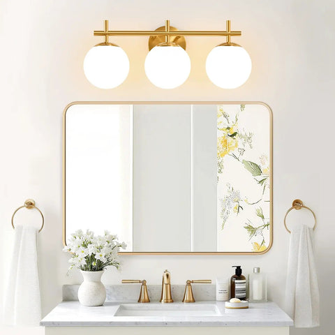 Next Level Three Light Bathroom Vanity Lights In Brushed Gold Finish With Milky White Glass Globe Shades Modern Wall Sconce Over Mirror above a stylish bathroom vanity with floral accents.