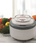 Digital Food Dehydrator With Smooth Adjustable Temperature And Even Heating For Whipping Up Homemade Beef Jerky Dried Fruit Fresh Herbs And Crispy Snacks On Four Stackable Trays In A Sleek Gray Design With Fresh Vegetables In Background.