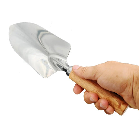 Hand holding Built To Last Stainless Steel Garden Shovel With Ergonomic Wooden Handle For Digging, Planting, Lawn Care And Trowel Tasks.