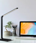 LED Desk Lamp With USB Port For Home Office And Reading, Eye Caring Light With 5 Color Modes And 5 Dimmable Levels, Touch Control And Auto Timer, Piano Style Modern Table Lamp next to laptop and plant on desk.