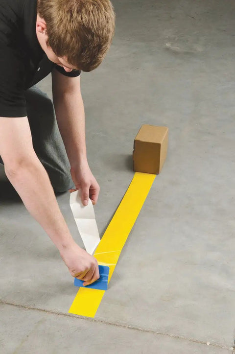 Heavy Duty Polyester Floor Marking Tape In Bright Yellow With Kick Ass Adhesion For Concrete Warehouses and 2 Inch By 100 Foot Roll being applied to a concrete floor.