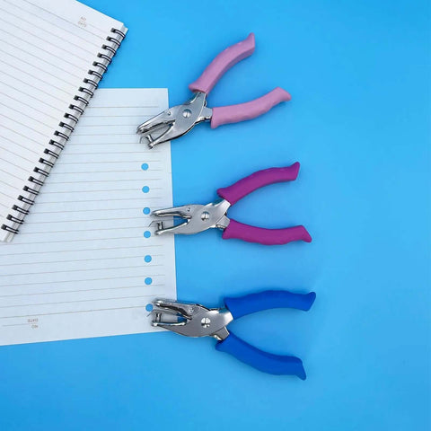 Three 1/4 Inch Hole Punchers for Crafts with soft grips in pink, purple, and blue, next to punched notebook paper on a blue background.