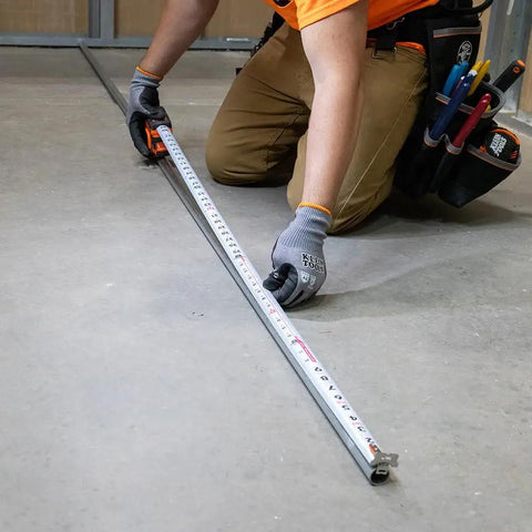 Worker using 9235 Heavy Duty Magnetic Tape Measure With 16 Foot Reach, showing nylon blade and double hook in action on concrete floor.