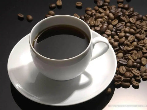 A cup of black coffee on a white saucer beside a pile of coffee beans on a black surface.