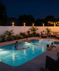 Outdoor nighttime scene with tiki torches lighting a pool area; people relaxing in water and seated on patio furniture near white fence.