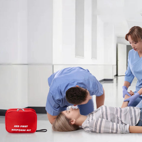 Red AED First Response Kit bag, front view, on the floor as two clinicians attend to a patient.