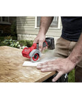 Milwaukee 12V cut-off tool, bare tool; red body with gray guard cutting a white rectangle on a wooden workbench (side view).