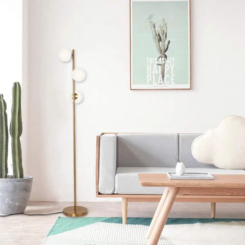 Three Globe Brushed Brass Floor Lamp illuminating a modern living room with a couch and cactus, creating a warm cozy vibe.