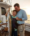 Father and son holding a sailfish on a boat at sunrise, with fishing rods in the background.