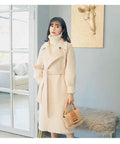 Woman in a cream coat with a belt, holding a handbag, standing in a stylish living room setting.