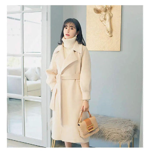 Woman in a cream coat with a belt, holding a handbag, standing in a stylish living room setting.