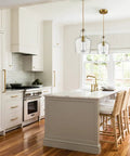 Lit Brushed Gold Pendant Light With Clear Glass Shade hanging over kitchen island with white cabinets and wood flooring