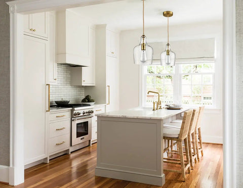 Lit Brushed Gold Pendant Light With Clear Glass Shade hanging over kitchen island with white cabinets and wood flooring