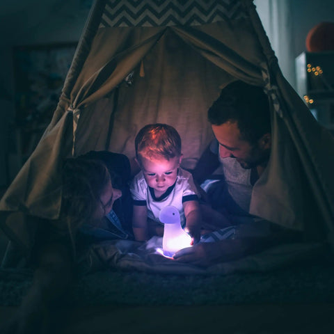 A family in a tent with a child holding a Multicolor LED Silicone Night Light Brontosaurus Tap Light For Kids Nursery Decor Rechargeable Color Changing Soft BPA Free Body Cozy Sleep Glow Bedtime Routine.