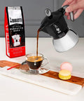 Heavy Duty Moka Induction Coffee Maker With Rock Solid Stainless Steel Boiler And Premium Aluminum Lid Brews Smooth Two Cup Espresso That Nails Your Morning Kickstart In Deep Black pouring espresso into a glass cup on a wooden and marble surface.