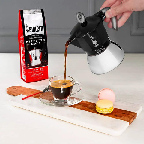 Heavy Duty Moka Induction Coffee Maker With Rock Solid Stainless Steel Boiler And Premium Aluminum Lid Brews Smooth Two Cup Espresso That Nails Your Morning Kickstart In Deep Black pouring espresso into a glass cup on a wooden and marble surface.