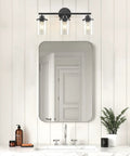 Three Light Matte Black Vanity Light With Clear Glass Shades For Bathroom above a mirror on a white paneled wall.