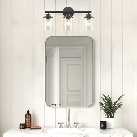 Three Light Matte Black Vanity Light With Clear Glass Shades For Bathroom above a mirror on a white paneled wall.