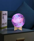 Colorful galaxy-themed night light on a wooden stand, emits purple and pink glow, beside books, on a bedside table.