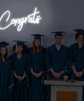 White Congrats Neon Sign Dimmable LED Wall Art For Graduation Party And Wedding Backdrop 17.7 By 9 Inches with graduates under it.