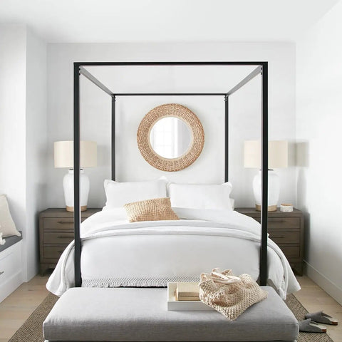 Minimalist bedroom with canopy bed, white bedding, wicker mirror, and two modern lamps on wooden nightstands.