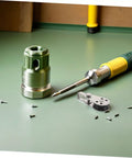 Close-up of a screwdriver, a metal adapter, and screws on a green surface with a yellow tool in the background.