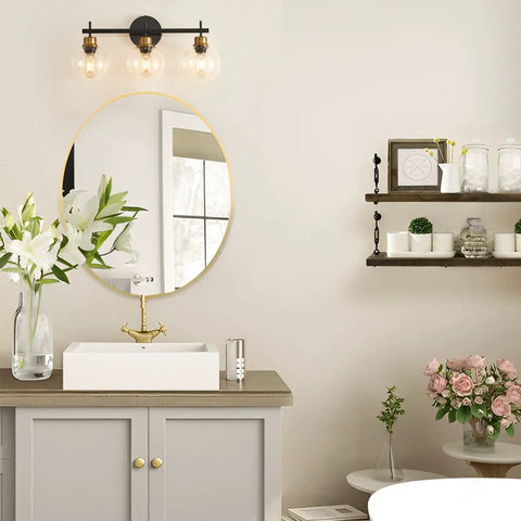 Three Light Black And Gold Bathroom Vanity Light Fixture With Metal Base And Clear Glass Globe Shades, Modern Over Mirror Bath Lighting That Elevates Your Space With Sleek Durable Build And High End Look installed above round mirror in bathroom setting.