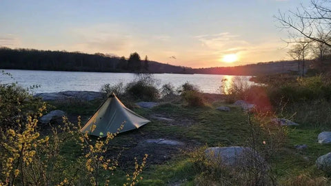 Gatewood Cape Ultralight 11 Oz Backpacking Tarp Shelter And Poncho Canopy set up by a lake at sunset, surrounded by trees.