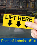 Front view: yellow Lift Here label decal, 5x2 in, with three down arrows, vinyl, held by hand (4-pack)