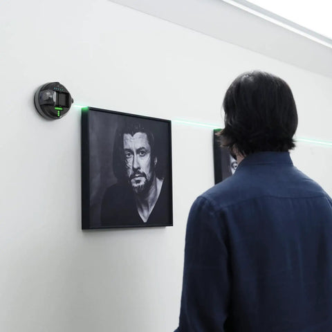 Person using MCube02 Laser Level Tool With Vertical And Horizontal Green Line Lasers on wall for picture hanging, 360-degree rotating base visible.