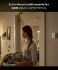 Person carrying laundry with wall-mounted motion sensor in home, Motion Sensor Bundle With Two White A19 LED Bulbs For Smart Home Lighting Hands Free Activation, Auto Day Night Adjustment, Customizable Brightness, App Control, Easy Setup.