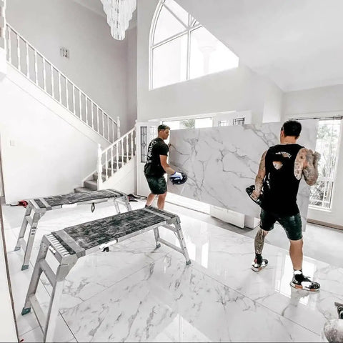 Two men lifting large marble slab with 8 Inch Electric Vacuum Suction Cup Heavy Duty Lifter For Glass Wood Drywall Granite Tile Sucker With One Button Start And Automatic Pump To Move Large Panels Up To 440 Lbs No Sweat in bright room. Ladder and stairs visible.