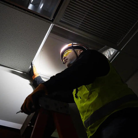 Electrician on ladder wearing Rechargeable Wide Beam Headlamp With Strap And 575 Lumens Five Lighting Modes All Day Runtime Direct Focus And Hard Hat Compatibility For Work And Outdoor Adventures, fixing ceiling panel.
