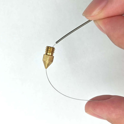 Hand holding a cleaning needle next to a brass 3D printer nozzle from the Complete 42Pcs 3D Printer Nozzle Cleaning Tool Kit.